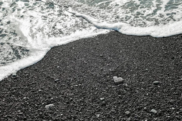 Uma pedra cinzenta em forma de coração encontra-se na costa com espuma do mar e água clara em uma praia de areia preta. Conceito de amor, casamento, liberdade. Fundo para cartão de Dia dos Namorados com espaço de cópia . — Fotografia de Stock