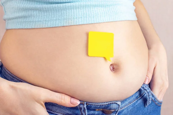 An empty yellow paper blank message on belly fat with hands squeezes at the waist of a young woman in jeans. The concept of excess weight, weight loss, diet, obesity, junk food. Mock up.