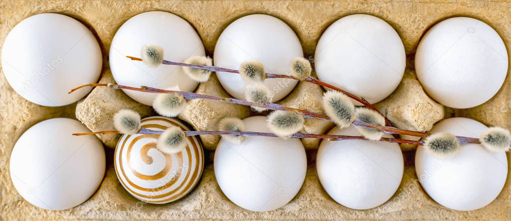 White unpainted chicken eggs in a cardboard box with willow branch close-up. One egg with a spiral pattern in gold paint. Easter traditional food, festive breakfast.