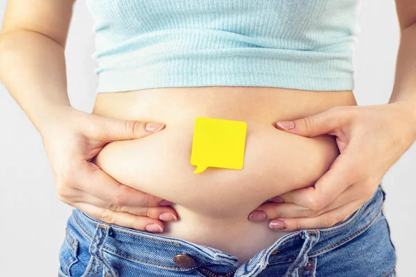 A yellow paper blank message is empty on belly with hands squeezes at the waist of a young woman in jeans. The concept of excess weight, weight loss, obesity, junk food.