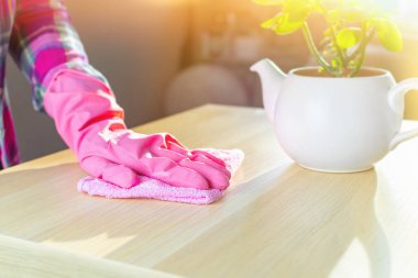 Pembe eldivenli bir kadının eli yumuşak bir bez tutarak masayı tozdan temizler. Ev temizliği, güneşli bir günde oturma odasındaki ışık yüzeylerini yıkamak, temizlik kavramı..