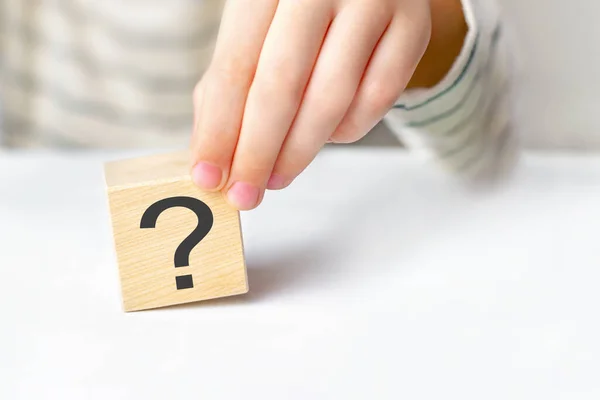 The concept of choice, decision making, solve a problem, find answer, learn the unknown. Close-up hand of a child holding the question mark on wooden block cube on white table background, copy space.