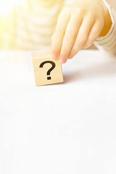 Close-up hand of a child holding the question mark on wooden block cube on white table background, sunny day, copy space. The concept of choice, decision making, solve a problem, find answer, unknown.