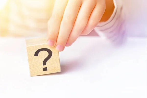 The concept of choice, decision making, solve a problem, find answer, unknown. Close-up hand of a child holding the question mark on wooden block cube on white table background, sunny day, copy space.