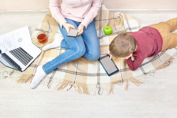 Genç bir kadın ve çocuk dairede yerde yatıyor. Anne bir telefon tutuyor, evden çalışıyor ve oğlu ekoseye uzanıyor ve tablet üzerinde oynuyor, uzaktan çalışma, serbest çalışma, çocuklar arasında çalışma, denge..