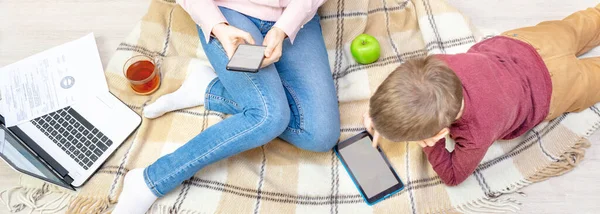 Ekoseli genç bir kadın ve çocuk. Anne bir telefon tutuyor, evden çalışıyor ve oğlu yakınlarda yatıyor ve tabletle oynuyor, uzaktan kumanda, serbest çalışma, çocuklar arasında çalışma, denge..
