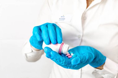 Doktor avucundaki ağrı ve sağlığı tedavi etmek için bir doz ilaç, vitamin ve antibiyotik veriyor. Mavi tıbbi eldivenli eller pembe ve beyaz haplarla dolu bir şişe tutuyor..