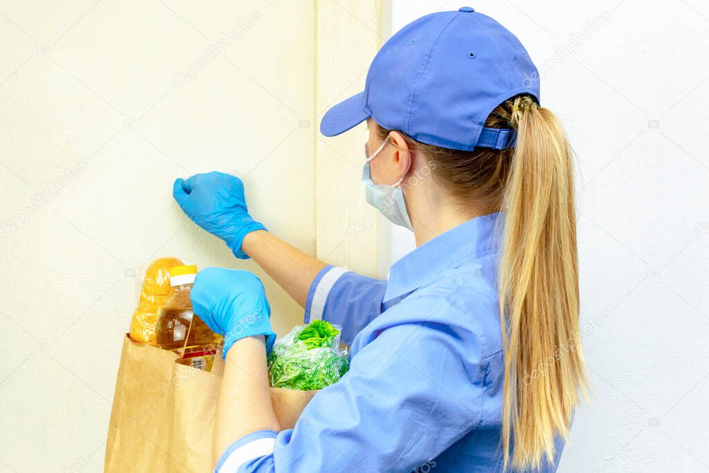 Donation, quarantine support, coronavirus. Volunteer young blonde woman in a uniform and a medical mask and gloves. Female knocks on the door and holds a paper bag with food. Delivery of purchases.