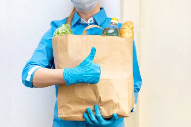 Mavi üniformalı, tıbbi maske ve eldiven takmış genç bir kadının gönüllüsü bir jest başparmağı gösteriyor, elinde sebzeli bir kese kağıdı tutuyor, yakın plan. Bağış, karantina, koronavirüs. Yemek teslimatı.