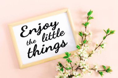 Üzerinde beyaz yazılar olan ahşap çerçeve küçük şeylerin tadını çıkarın, bahar ilham verici ve motive edici metinler ve yumuşak pembe arka planda çiçek açan çiçek açan kiraz dalları. Üst görünüm.