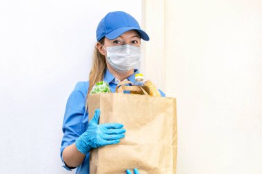Mavi üniformalı ve şapkalı, tıbbi maske takmış ve eldiven takmış genç bir kadının portresi elinde yiyecek ve sebzelerle dolu bir kese kağıdı tutuyor. Bağış, karantina ve koronavirüsteki insanlara yardım.