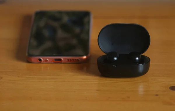 Smartphone and wireless black headphones in a charging case on an oak table