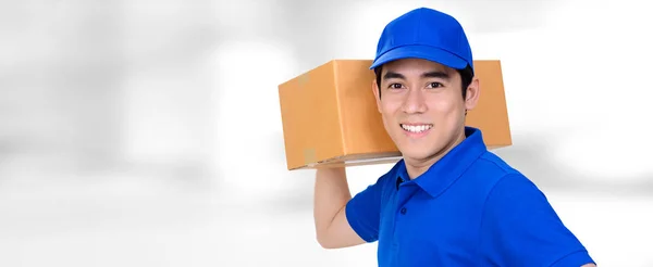 Smiling friendly delivery man carrying parcel box on blur panormic white background — Stock Photo, Image