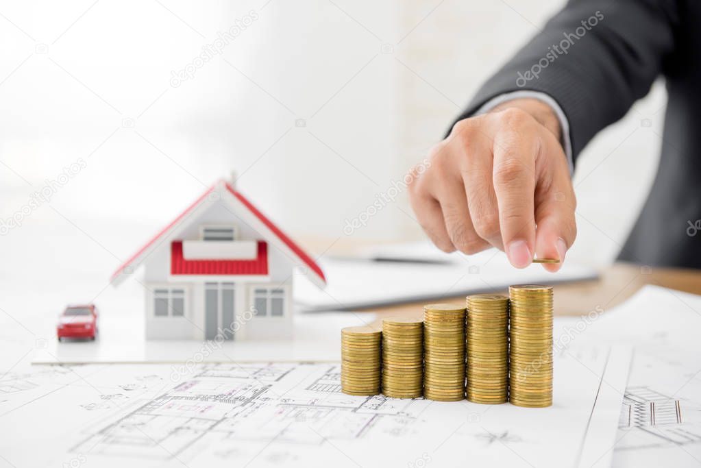Businessman putting coin on top of money stacks with blurred house model in background - real estate financial and investment concepts