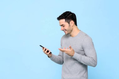 Gülümseyen mutlu genç adam açık mavi stüdyo arka planında izole akıllı telefon kullanıyor.