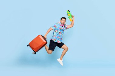 Songkran festivali sırasında su tabancası ve bagaj taşıyan yakışıklı Asyalı turist mavi arka plan çekti.