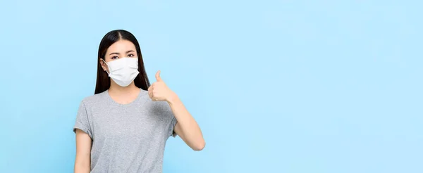 年轻的亚洲女人戴着医疗面罩 无声无息地把大拇指放在浅蓝色横幅背景下 并留有复制空间 — 图库照片