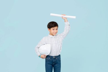 Yakışıklı, gülümseyen, kasklı, geleceğin mühendisi olmak isteyen Asyalı çocuk açık mavi stüdyo arka planında izole edilmiş planlarını gösteriyor.
