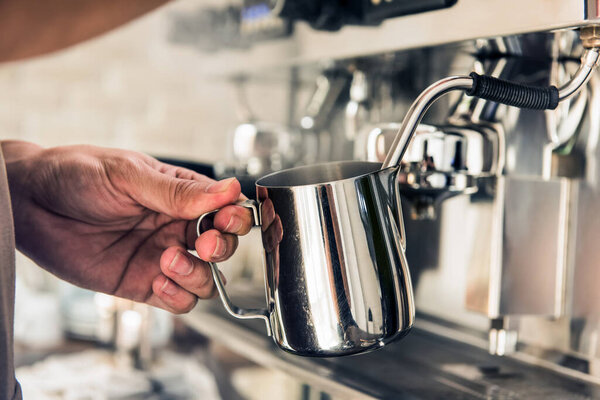 Barista steaming milk in the pitcher with coffee machine preparing to make latte art in cafe