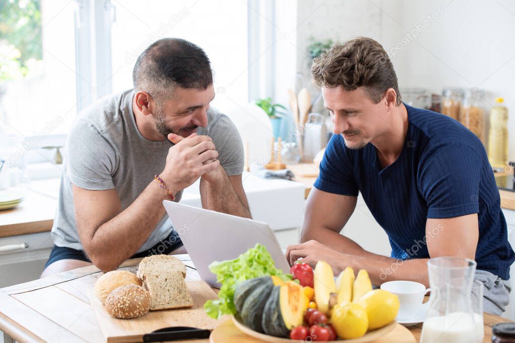 Fit mature male caucasian gay couple having a happy morning conversation while working with a laptop in the kitchen