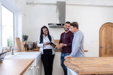 African american female real estate agent in kitchen showing gay couple around new house clipart