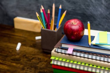 Renkli kitap yığını, kırtasiye ve eğitim malzemeleri sınıfın ahşap masasında arka planda tahtayla birlikte.