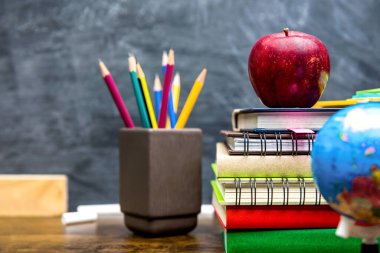 Renkli kitap yığını, kırtasiye ve eğitim malzemeleri sınıfın ahşap masasında arka planda tahtayla birlikte.