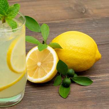 Limonata suyu bardakta tazeleniyor, ahşap masa arkasında limonlar var.
