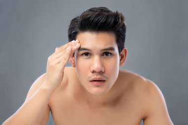 Yüzünü cilt bakımı ve güzellik konseptleri için kontrol eden tişörtsüz genç Asyalı adamın portresi, gri arka planda izole edilmiş stüdyo çekimi.