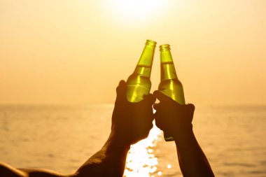 Bira şişelerini tutan bir çift el yaz günbatımında sahilde tatil yapıyor ve kutluyor.