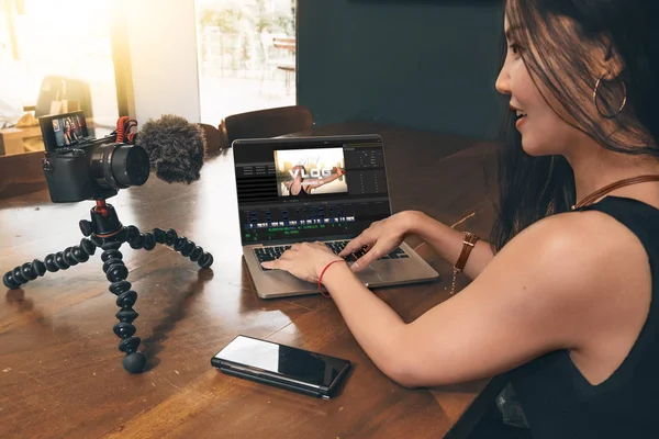 Asian woman editing vlog film on a laptop. Camera and phone on the table.