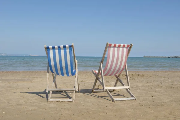 Dwa puste płótno plaży krzesła, jeden niebieski i jeden czerwony w środku obrazu na plaży Weymouth w Wielkiej Brytanii, widokiem na morze w słoneczny dzień — Zdjęcie stockowe
