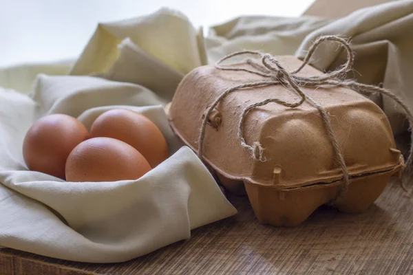 Delicate still life with three brown eggs on a light cloth as a nest and a egg carton packed with string on a kitchen board. Soft beige tones.Horizontal view.