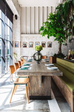 Yeşil kanepe, ahşap masa, tahta sandalyeler, yeşil bitki ve beyaz çizik desenli duvar kağıdı ile dekore edilmiş grup yemek masası. Modern restoran atmosferi.