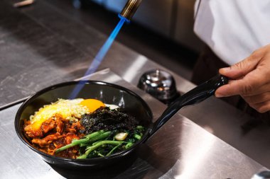 Bibimbap (Kore pilavı kimchi, yosun ve susamlı kızarmış sebzeyle karıştırılmış) sıcak tavada servis edilen şefin kızarmış yumurtanın üzerinde mozzarella peyniri eritmek için kaynak makinesi kullanması..