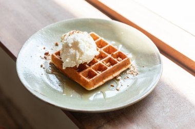 Bir kepçe vanilyalı dondurmayla ev yapımı klasik waffle. Ezilmiş badem ve akçaağaç şurubuyla kaplanmış. Seramik tabakta servis edilir..