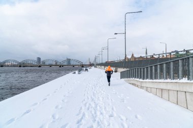 Sporcu kışın koşar ve traktör kar sürer. Fotoğraf arkadan Riga