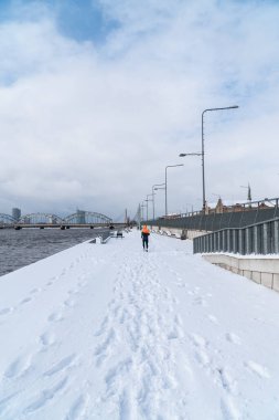 Sporcu kışın koşar ve traktör kar sürer. Fotoğraf arkadan Riga