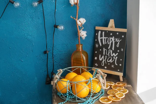 whole and cut oranges on the table, a vase with a twig and a Board with the inscription happy new year on the nightstand against the blue wall with a garland
