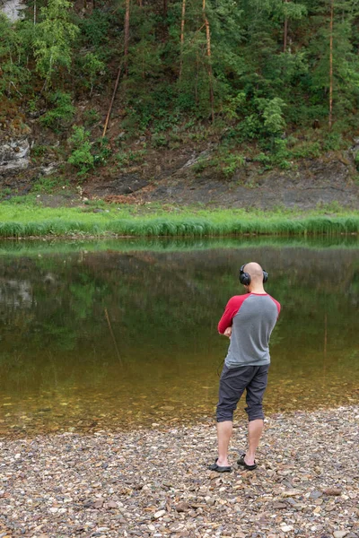 Back view of young hipster man listening to music in headphones on river bank. Copy space. Far from big city bustle. Relaxing outdoors. Tourism, active leisure. Best company