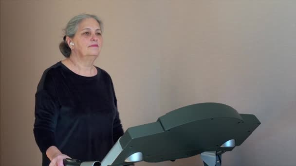 Sporty senior woman is training on treadmill in home and looking in mobile phone. Checking social media. Received an email. — Stock video