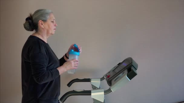 Mulher sênior está bebendo água de garrafa shaker enquanto caminha na esteira — Vídeo de Stock