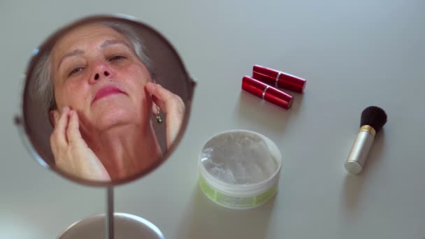 거울에 만진 얼굴을 하고 있는 성숙 한 여성. 흰 상 위에 화장품을 놓음 — 비디오