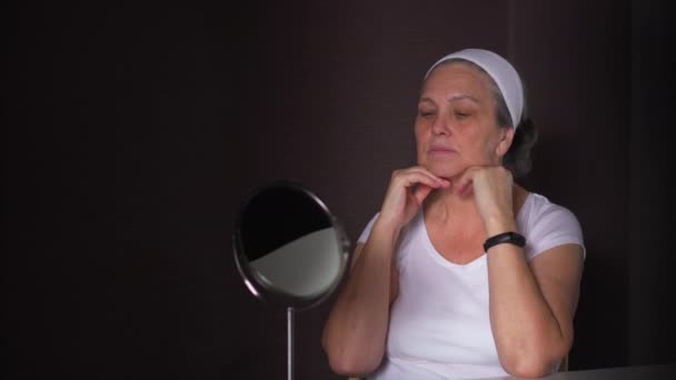 Aged woman with white headband looking in mirror aging process of skin — Stock Video