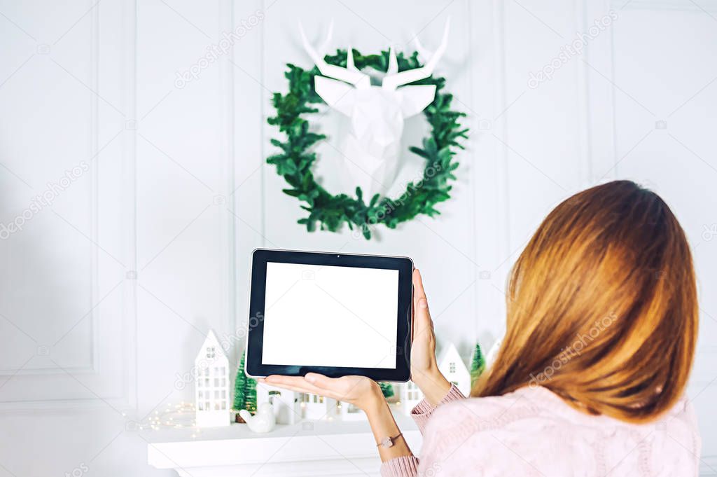 Mock up of the tablet in the hands of the girl. On the background of the wall with a decorative deer and a wreath with houses of festive.