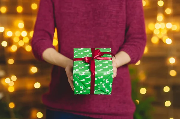Una ragazza con un maglione bordeaux, tiene tra le mani un regalo di scatola verde con un nastro rosso. Sullo sfondo una parete di legno con una ghirlanda di stelle. Concetto sul tema delle vacanze . — Foto Stock