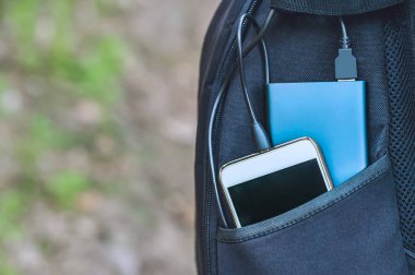 Bir güç bankası akıllı telefonu otların ve kumların arasında siyah bir sırt çantasının cebinde şarj eder..