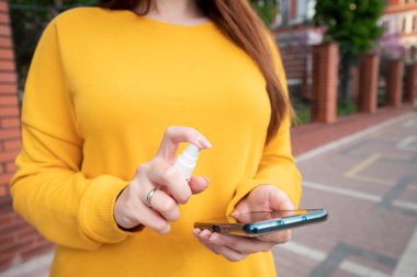 Sarı kazaklı genç bir kız telefonunu sprey dezenfektanla dezenfekte ediyor.
