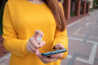 Sarı kazaklı genç bir kız telefonunu sprey dezenfektanla dezenfekte ediyor.