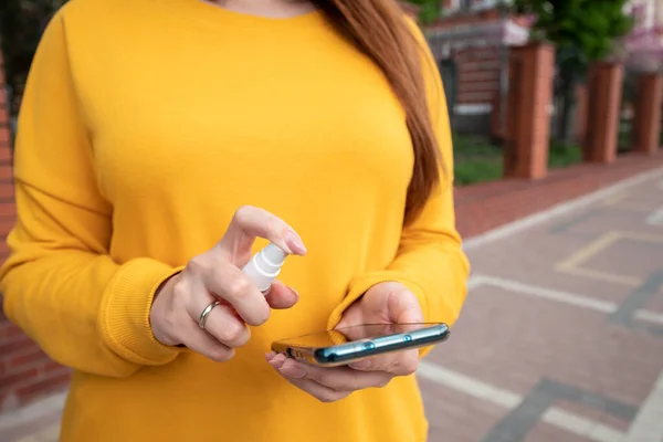Sarı kazaklı genç bir kız telefonunu sprey dezenfektanla dezenfekte ediyor.